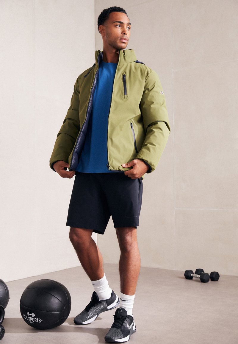 Chaqueta aislante verde oliva con cuello alto, bolsillos con cremallera, llevada sobre una camisa azul y pantalones cortos negros, sobre un fondo neutro.