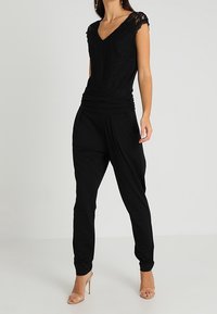 Black lace top with a V-neck and short sleeves, paired with loose-fitting black trousers. Worn with clear high-heeled shoes.