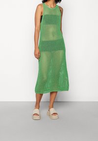 Abito in rete verde con design senza maniche, lunghezza al ginocchio, caratterizzato da una trama a maglia aperta. Abbinato a sandali slip-on nei toni neutri.
