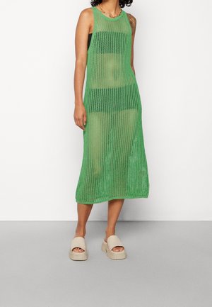Grünes Netzkleid mit einem ärmellosen Design, knielang, mit einer offenen Strickstruktur. Dazu passende neutrale Schlupfsandalen.