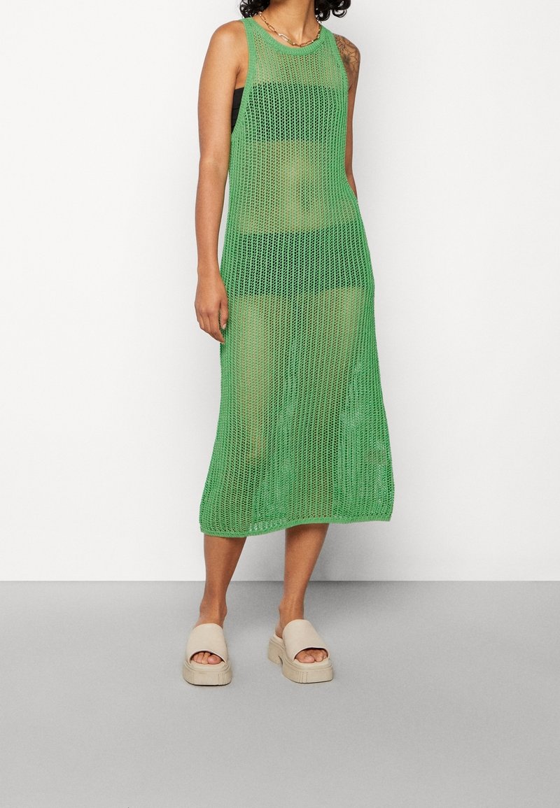 Abito in rete verde con design senza maniche, lunghezza al ginocchio, caratterizzato da una trama a maglia aperta. Abbinato a sandali slip-on nei toni neutri.