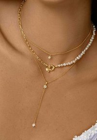 Collares de tres capas: una cadena de oro, una con perlas y una cadena de oro con acentos de estrellas. Todos tienen un acabado pulido.