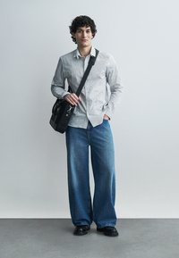 Νέος άνδρας με σγουρά μαλλιά, που φοράει πουκάμισο με σχέδιο, μπλε τζιν παντελόνι με φαρδιά μπατζάκια, μαύρα παπούτσια και κρατάει μαύρη τσάντα ώμου.