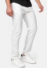 Mand iført hvidt slim-fit jeans og hvide sneakers med sorte detaljer, stående mod en ensfarvet baggrund.