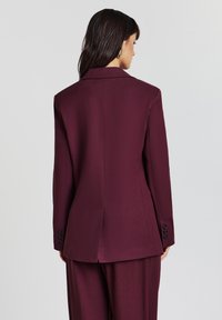 Una donna con lunghi capelli scuri indossa un blazer bordeaux su misura e pantaloni abbinati, vista di spalle su uno sfondo chiaro e semplice.