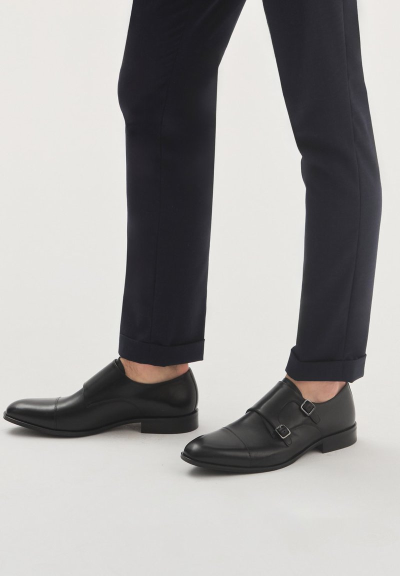 Czarne skórzane buty do garnituru z podwójnymi zapięciami; elegancki design i gładka tekstura; zestawione z granatowymi spodniami, skróconymi na kostkach.