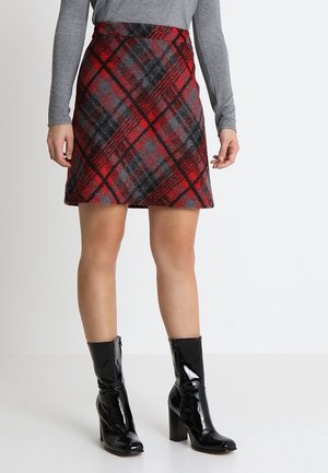 Femme portant une jupe à carreaux rouge et noire, un haut gris à manches longues et des bottes noires en cuir verni à talons mi-mollet, debout sur un sol blanc.