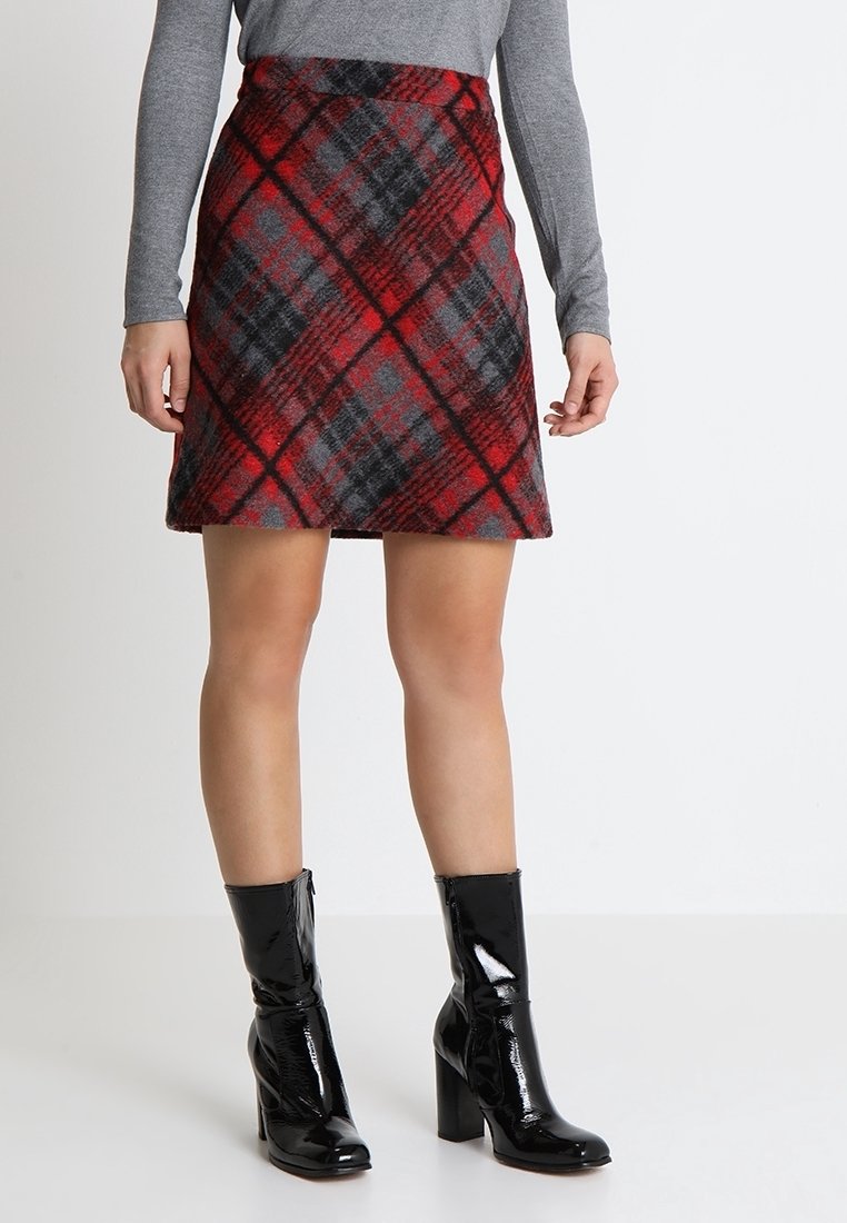 Femme portant une jupe à carreaux rouge et noire, un haut gris à manches longues et des bottes noires en cuir verni à talons mi-mollet, debout sur un sol blanc.