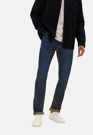 Mężczyzna ubrany w ciemnoniebieskie dżinsy z mankietami przy kostkach, białe tenisówki, białą koszulkę i czarną zapinaną na guziki kurtkę, stojący z jedną ręką w kieszeni.