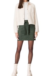 Mujer vistiendo una chaqueta de forro polar blanca y corta sobre una blusa blanca, falda verde de cintura alta con lazo frontal, medias negras transparentes y botas blancas hasta el tobillo.