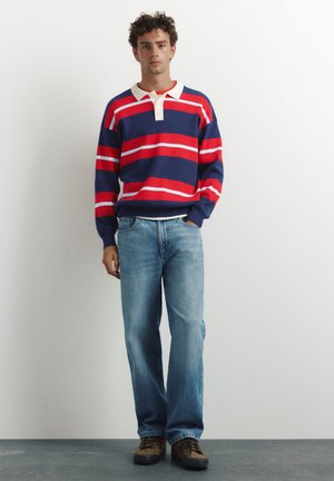 Jeune homme debout portant un maillot de rugby rayé bleu marine, rouge et blanc, un jean bleu clair et des chaussures marron à lacets, devant un mur blanc uni.