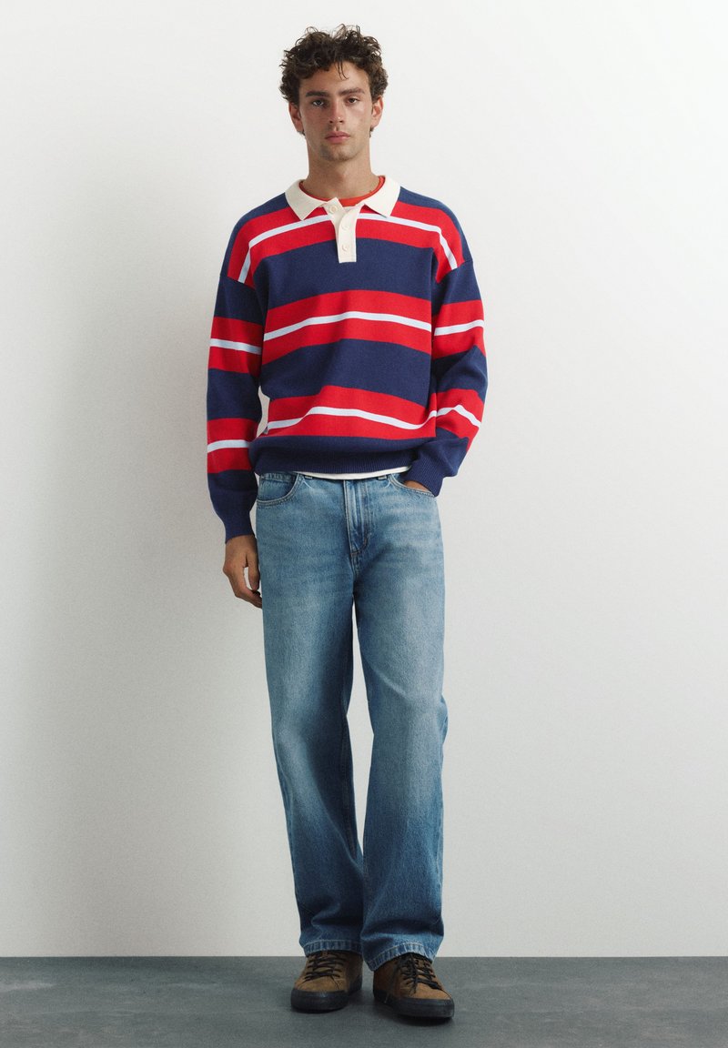 Jeune homme debout portant un maillot de rugby rayé bleu marine, rouge et blanc, un jean bleu clair et des chaussures marron à lacets, devant un mur blanc uni.