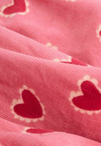 Rosa tyg med ribbad textur, prytt med upprepade röda hjärtan som är omgivna av vitt. Hjärtana är oregelbundet placerade över materialet.