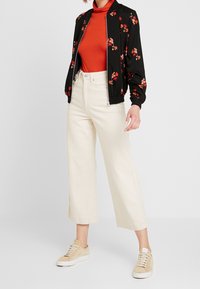 Bomber jacket nera con stampa floreale, dolcevita rossa, pantaloni beige a gamba larga e sneakers chiare. Tessuto morbido e vestibilità rilassata.