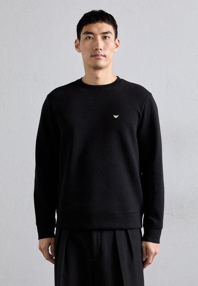 Sweatshirt noir en tissu lisse, avec un col rond et des manches longues. Un petit accent logo doré se trouve sur la poitrine gauche.