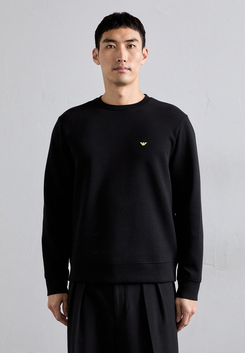 Black sweatshirt made from a smooth fabric, featuring a round neckline and long sleeves. A small gold logo accent is on the left chest.