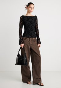Črna dolga majica z cvetličnim čipkastim detajlom, rjave široke hlače in črna usnjena torba čez ramo. Model nosi črne sandale z zaponkami.