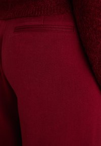 Red fabric trousers with a smooth texture; features a back pocket with a double stitched trim, alongside a fuzzy red sweater overlay.