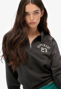 Felpa nera con zip caratterizzata da ricamo bianco "Superdry 23", con colletto alto e morbida texture del tessuto.