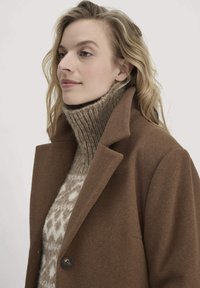 Brauner Wollmantel mit fallenden Revers und ein-Knopf-Verschluss, darüber ein beiger Rollkragenpullover mit Muster. Glatte Textur und klassischer Schnitt.