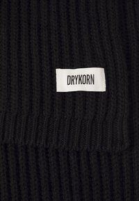 Sort ribbet garn med en hvid rektangulær label syet på, der viser mærkenavnet "DRYKORN" i sorte versaler.