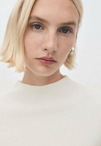 Femme blonde avec les cheveux courts, yeux bleus, légères taches de rousseur, portant un haut blanc et des boucles d'oreilles en argent, regardant directement l'objectif.