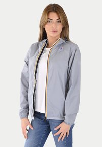 Giacca a vento grigio chiaro con polsini elastici, cerniera frontale e dettagli colorati a righe lungo la cerniera. Indossata sopra una maglietta bianca.