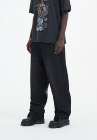 Černé grafické tričko s barevnými designy, v kombinaci s volnými černými džíny, které mají na boku složitou výšivku, a robustními černými botami.