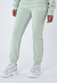 Sudadera verde menta con un ajuste slim, que presenta puños en los tobillos, bolsillos laterales y un logo blanco en el muslo izquierdo. Combinada con zapatillas blancas de suela gruesa.