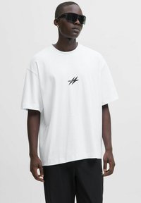 Mand iført en hvid oversized t-shirt med et lille sort logo, sorte bukser, store sorte solbriller og en lille hoops-ørering, der står mod en hvid baggrund.