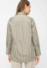 Camicia a righe verde oliva e bianca, caratterizzata da un colletto, maniche lunghe con polsini a bottoni e una schiena plissettata per una maggiore libertà di movimento.