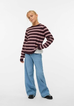 Suéter a rayas en púrpura oscuro y rosa claro, de corte relajado, combinado con unos jeans de pierna ancha en azul y calzado negro, de pie sobre un fondo blanco.