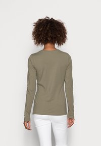 Person mit lockigem Haar, der ein olivgrünes Langarmshirt und weiße Hosen trägt, von hinten vor einem schlichten Hintergrund gesehen.