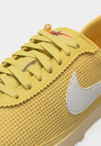 Κίτρινο αθλητικό παπούτσι Nike με υφασμάτινο άνω μέρος με υφή, κίτρινες κορδόνια που ταιριάζουν, λευκό λογότυπο Nike swoosh και μερικώς ορατό πορτοκαλί λογότυπο στη γλώσσα.