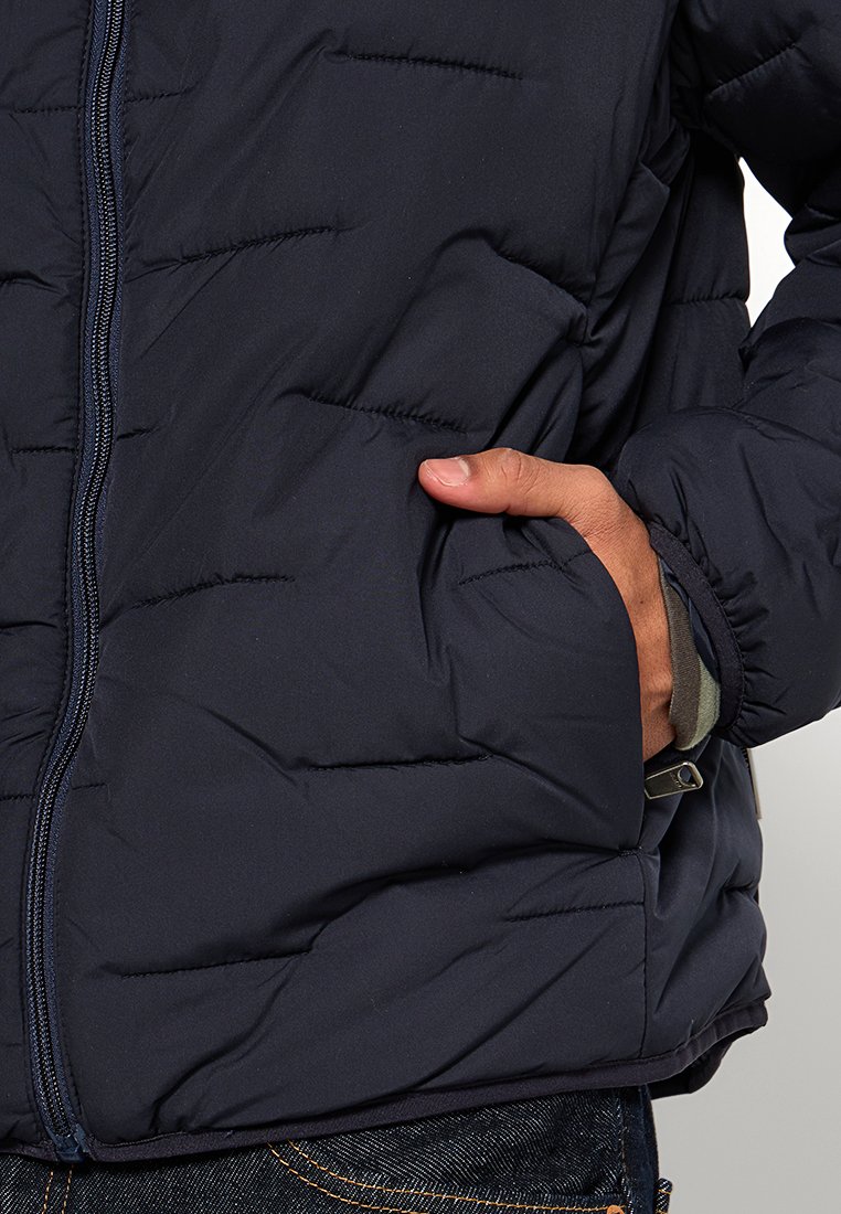 Marineblaue gepolsterte Jacke mit Reißverschluss vorne, ausgestattet mit einer strukturierten Oberfläche und Seitentaschen. Die Bündchen sind elastisch für einen eng anliegenden Sitz.