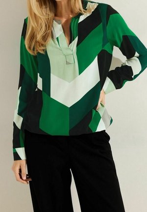 Vrouw draagt een blouse met lange mouwen met geometrische groene, witte en zwarte patronen en een zwarte broek, staand met één hand op haar heup.