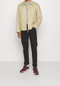 Light beige jacket with a zipper and two pockets, grey sweater underneath, black pants, and maroon running shoes with white soles.