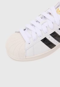 Sneaker de couro branco com biqueira de borracha, atacadores brancos e riscas pretas. Apresenta uma biqueira texturizada e detalhes costurados.
