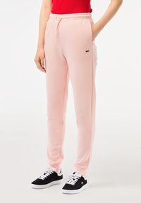 Pantalones de chándal rosas con cintura y puños elásticos, con un pequeño logo verde. Combinados con zapatillas negras y blancas. Textura suave.