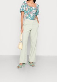 Blusa floreale teal con maniche corte e scollatura quadrata, abbinata a pantaloni larghi verde chiaro. Gli accessori includono una piccola borsa gialla e sandali verde con tacco.