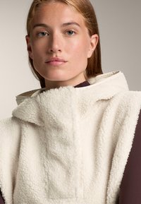 Crème-kleurige fleece hoodie met een hoge hals, met een zachte textuur en capuchon. Draagt over een donkere top en laat een gelaagde look zien.