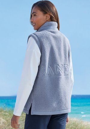 Frau steht im Freien am Meer, trägt eine hellblaue Fleece-Weste über einem weißen Langarmshirt und navyblaue Hose, lächelt und schaut nach links.