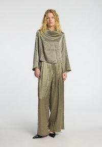 Olive green, textured top with a wide neckline and cropped fit, paired with loose, high-waisted trousers. Black, pointed shoes complete the look.