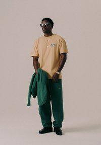 T-shirt graphique beige avec des détails en camouflage vert, associé à un pantalon de survêtement vert et un sweat à capuche coordonné, chaussures noires et lunettes de soleil carrées oversize.