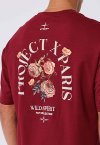Camiseta marrón por detrás con diseño floral y texto "PROJECT X PARIS", "WILD SPIRIT" y "PXP COLLECTION" debajo de las flores y el logo.