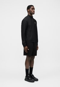 Melna sporta jaka ar augstu apkakli un rāvējslēdzēju, ar teksturētu audumu un sānu kabatām, kopā ar melniem šortiem un sporta apaviem.