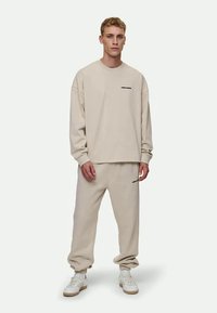 Beige Sweatshirt und passende Sweatpants aus weichem Material. Der Sweatshirt hat ein kleines schwarzes Logo; die Pants verfügen über ein ähnliches Logo und einen lässigen Schnitt.