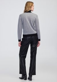 Pull gris avec des accents noirs, présentant un col montant, des manches longues et des poignets côtelés, associé à un pantalon en cuir noir, se tenant sur un fond uni.