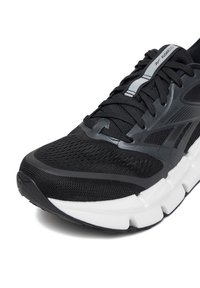 Čierna športová obuv s textilným zvrškom z oka, textúrovaným dizajnom a bielou podrážkou. Obsahuje čierne šnúrky a sivé akcenty na špičke a logu.