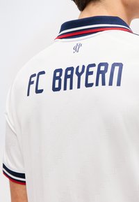 Άτομο που φοράει λευκό μπλουζάκι της FC Bayern με γιακά και διακοσμητικές ρίγες στα μανίκια σε ναυτικό μπλε, κόκκινο και λευκό, από πίσω.